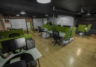Un espace de coworking chaleureux chez Nuage B à Montréal avec des panneaux de séparation verts, des chaises ergonomiques et un mélange de bureaux ouverts et semi-privés dans un cadre industriel.
